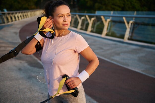 Mulher atlética determinada confiante, colocando fones de ouvido enquanto se exercita com tiras durante o treinamento com peso corporal em suspensão em uma ponte da cidade ao pôr do sol