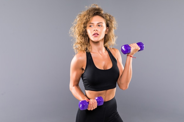 Mulher atlética fazendo exercícios para os braços. Foto do modelo de aptidão muscular malhando com halteres na parede cinza. Força e motivação