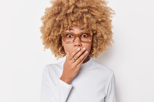 Mulher atônita com cabelo encaracolado cobre a boca com a mão usa óculos e poses de jumper casual contra fundo branco com medo de algo horrível. Conceito de reações e emoções humanas.