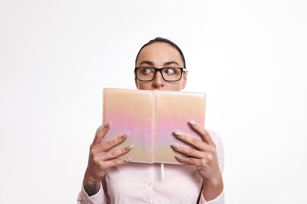 Mulher atordoada e surpresa olhando para longe e cobrindo a boca com um livro rosa. Isolado no fundo branco com espaço de cópia
