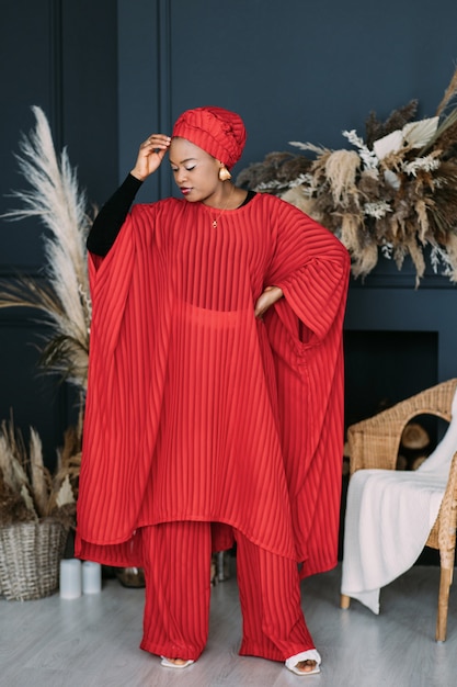 Mulher atraente afro-americana fazendo diferentes expressões emocionais em um elegante terno vermelho feito à mão e lenço na cabeça em um estúdio moderno com decoração rústica