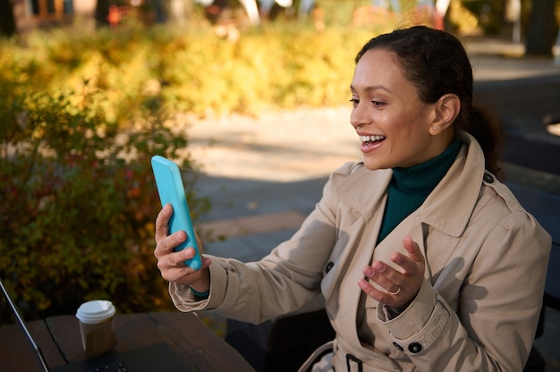 Mulher atraente alegre sorri ao fazer uma videochamada através de um aplicativo móvel em seu smartphone. Jovem mulher de negócios confiante trabalhando ao ar livre, longe da civilização urbana, em um café de outono no bosque de carvalho