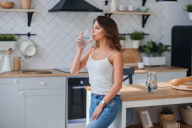 Mulher atraente bebe água pura na cozinha, de manhã. Conceito de cuidados de saúde e dieta.