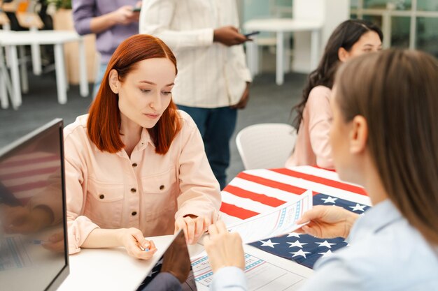 Foto mulher atraente com cabelo vermelho falando com um trabalhador no posto de votação sentado na mesa de registro