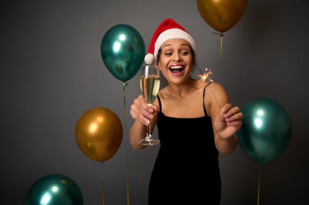 Mulher atraente com um lindo sorriso em um vestido de noite preto e chapéu de Papai Noel gosta de festa de Natal, segura taça de champanhe e luzes de Bengala, regozija-se com um fundo cinza com bolas de ar purpurina festivas
