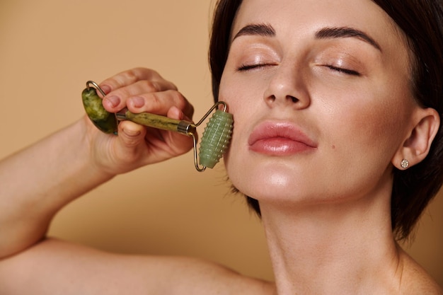 Mulher atraente de beleza natural massageia seu rosto com o rolo de pedra. Jovem morena caucasiana, fazendo massagem facial anti-envelhecimento, suavização e drenagem linfática com massageador de rolo de jade. Cuidados com a pele.