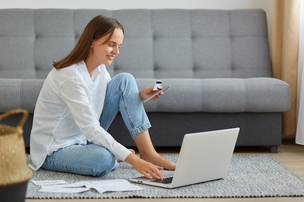 Mulher atraente de cabelos escuros vestindo camisa branca e jeans, segurando o cartão de crédito nas mãos, usando computador portátil para inserir dados, reservar tour ou fazer compras online, pagamento.