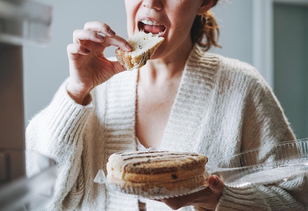 Mulher atraente de meia-idade no casaco de lã aconchegante comendo bolo da geladeira na cozinha em casa