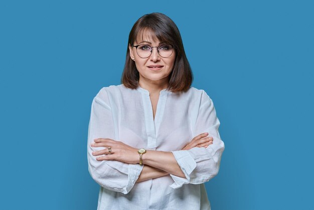 Foto mulher atraente de meia idade olhando para a câmera no fundo de cor azul sorrindo confiante mulher de 40 anos com óculos de braços cruzados em camisa branca estilo de vida confiança calma conceito de pessoas maduras