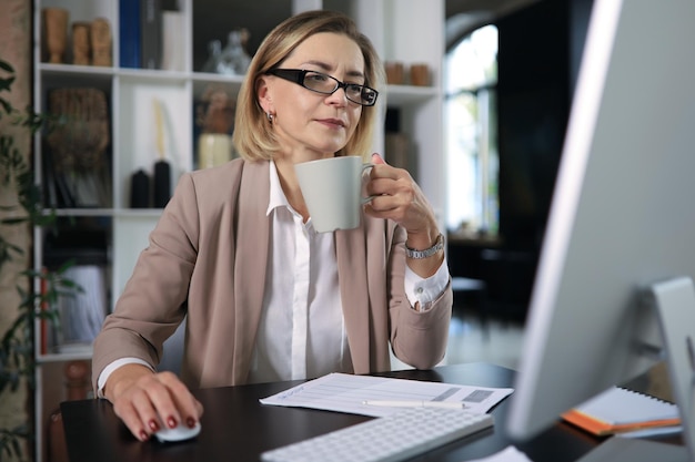 Mulher atraente de meia idade, trabalhando no escritório, usando o computador desktop contemporâneo.