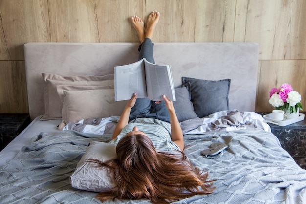 Mulher atraente deitada na cama pela manhã, bebendo café, lendo um livro, estilo casual, vestido cinza, sentindo-se confortável em casa, descansando, sorrindo