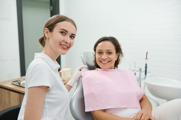 Mulher atraente e confiante, dentista higienista fofamente sorri olhando para a câmera ao lado da paciente sentada na cadeira do dentista antes de receber tratamento odontológico na clínica odontológica. Cuidados de higiene bucal