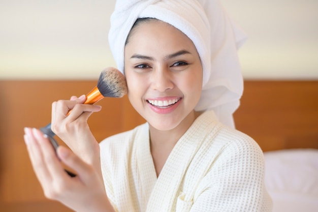 Mulher atraente e feliz em roupão branco está aplicando maquiagem natural com pincel de pó cosmético, conceito de beleza.