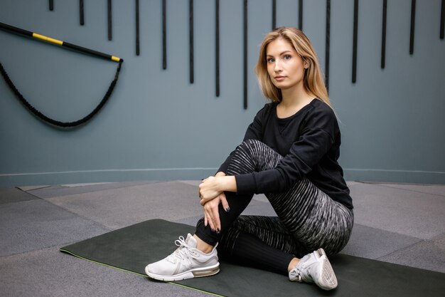 Mulher atraente e feliz sentada no chão após o exercício no fundo do ginásio. conceito de treino de menina magro e saudável.