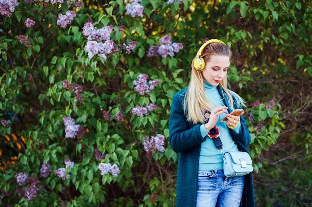 Mulher atraente é ouvir música e digitar em seu telefone ao ar livre no parque entre as flores lilás.