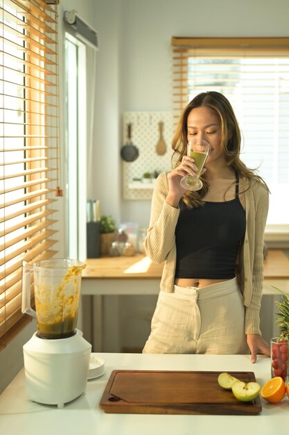 Mulher atraente em pé na cozinha e bebendo smoothie de desintoxicação verde caseiro Refeição vegana e conceito de estilo de vida saudável