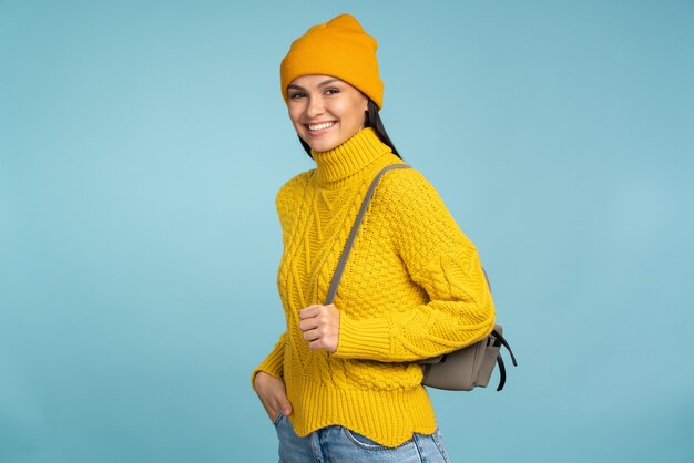 Mulher atraente em um elegante suéter amarelo sorri amplamente enquanto posava com sua mochila. Garota legal em roupas de malha quentes, em pé sobre o fundo azul. Conceito de inverno