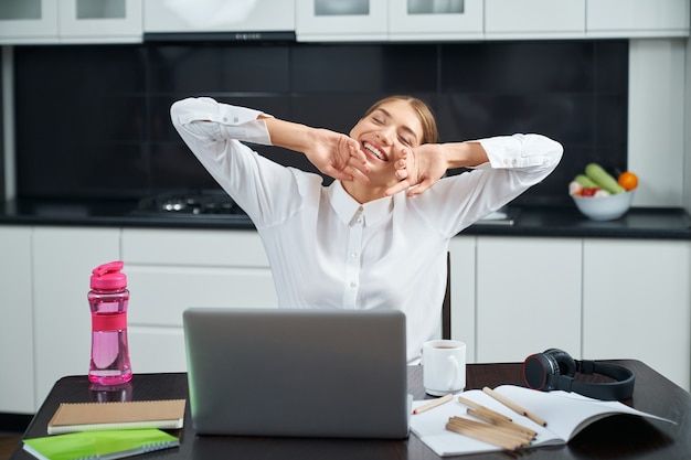 Mulher atraente esticando o corpo enquanto está sentado à mesa da cozinha durante o trabalho remoto no laptop