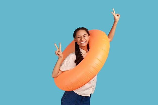 Mulher atraente feliz animada com dreadlocks pretos segurando o anel de borracha laranja quer piscina mostrando sinal de v para a câmera vestindo camisa branca Foto de estúdio interior isolada em fundo azul