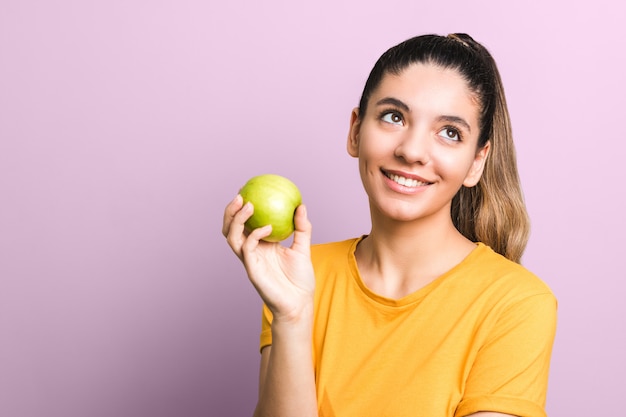 Mulher atraente jovem pensativa em camiseta amarela segurando a maçã verde e sorrindo pensando em comida lixo