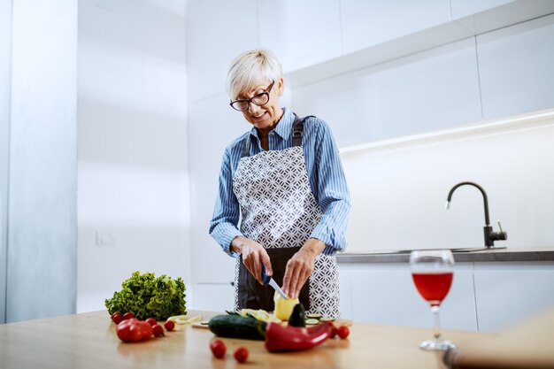 Mulher atraente loira sênior no avental em pé na cozinha em casa e cortar o pimentão amarelo