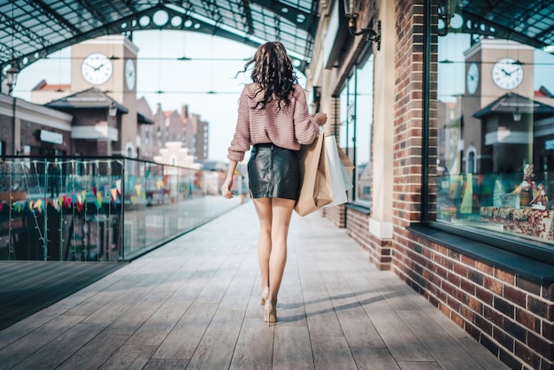 Mulher atraente morena de pernas compridas usando saia curta de couro e salto alto caminhando por um grande shopping center segurando um monte de sacolas de papel