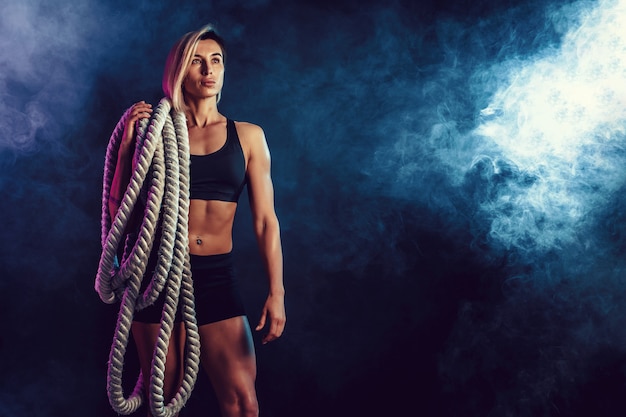 Foto mulher atraente no sportswear preto com cordas pesadas nos ombros na parede escura. força e motivação. desportiva mulher trabalhando com cordas pesadas.