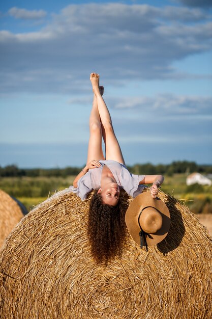 Mulher atraente posando nos campos de trigo ceifados com feixe e céu na aldeia. Fardos de feno no campo após a colheita