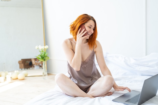 Mulher atraente ruiva de meia-idade de pijama, sentada na cama, usando telefone e laptop