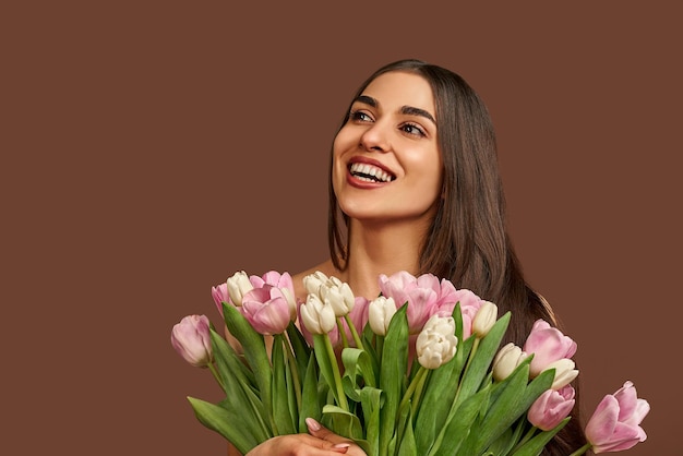 Foto mulher atraente segurando buquê de tulipas brancas e rosa em um fundo marrom ri e olha para cima