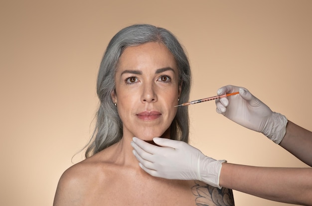 Mulher atraente sênior de cabelos grisalhos recebendo injeção cosmética em dobras nasolabiais em pé em bege