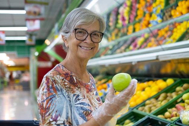 Mulher atraente sênior no supermercado, escolhendo frutas frescas, segurando uma maçã verde na mão. Frutas coloridas misturadas no fundo