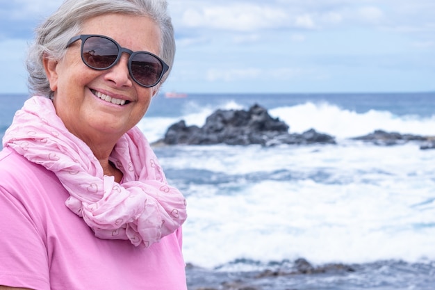 Mulher atraente sênior rosa vestida sorrisos na praia em dia de vento. Horizonte sobre a água