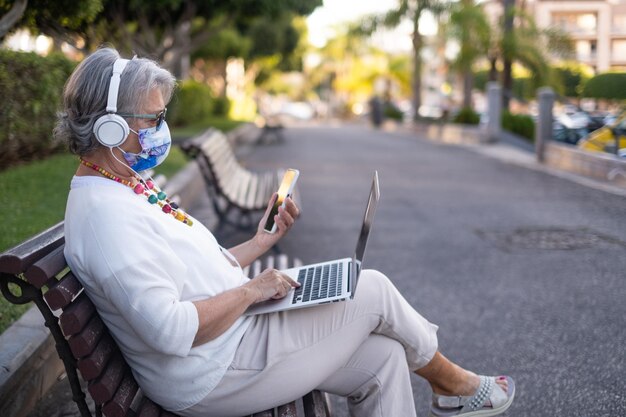Mulher atraente sênior sentada em um banco de parque usando telefone celular e laptop, aproveitando a tecnologia