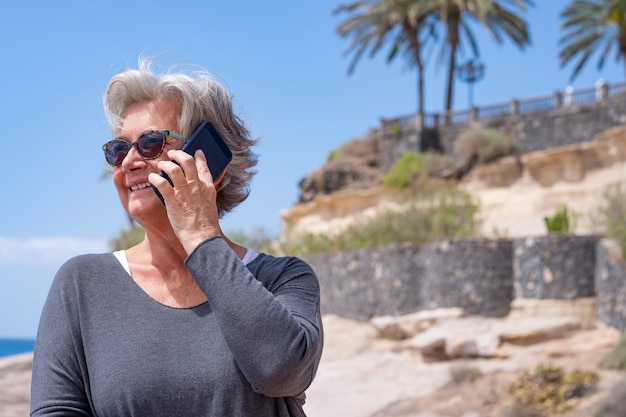 Foto mulher atraente sênior usando smartphone ao ar livre no mar sorrindo feliz