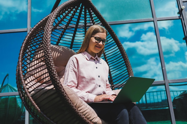 Mulher atraente senta-se com um laptop na varanda e trabalha olhando atentamente para a tela Uma linda garota freelancer em roupas casuais trabalha em um laptop enquanto está sentado em uma cadeira na varanda