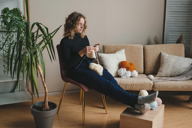 Mulher atraente sentada e relaxando em uma cadeira confortável em casa no fim de semana curtindo seus hobbies tricotando um cachecol um suéter Pessoas estilo de vida Estilo moderno Móveis modernos Estilo simples