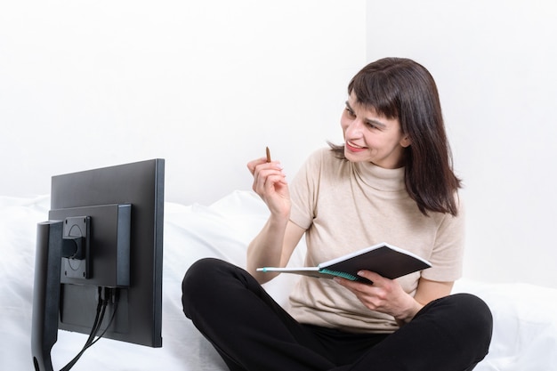 Mulher atraente sentada no sofá segurando um caderno e uma caneta e olhando atentamente para o monitor do computador em casa