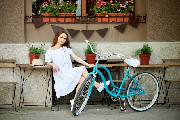 Mulher atraente, sentado do lado de fora na zona de café aconchegante com sua bicicleta azul vintage perto na frente do prédio decorado da cidade durante o dia quente de verão