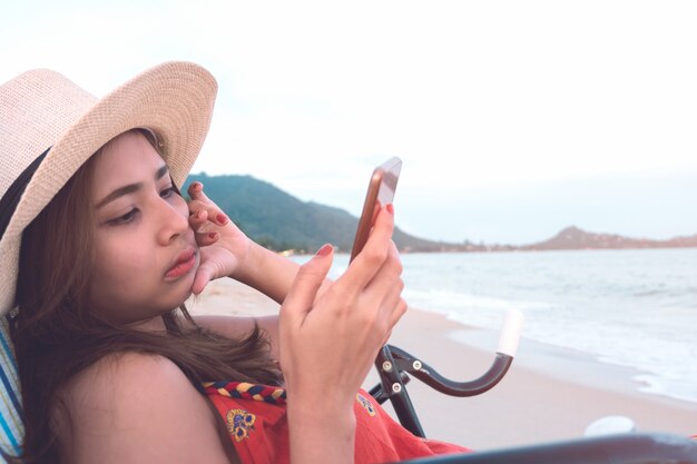 Foto mulher atrativa no chapéu usando o smartphone ao sentar-se na cadeira de praia na praia do por do sol.