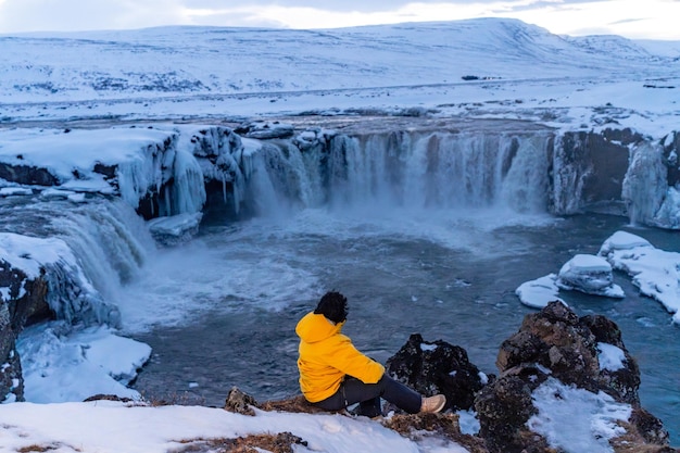 Mulher aventureira no inverno na Islândia sentada olhando para a cachoeira congelada de Godafoss