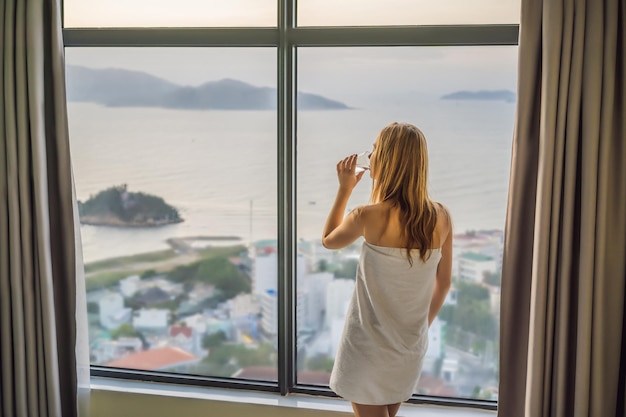 Mulher bebe água pela manhã no fundo de uma janela com vista para o mar