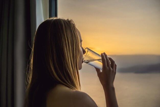 Mulher bebe água pela manhã no fundo de uma janela com vista para o mar