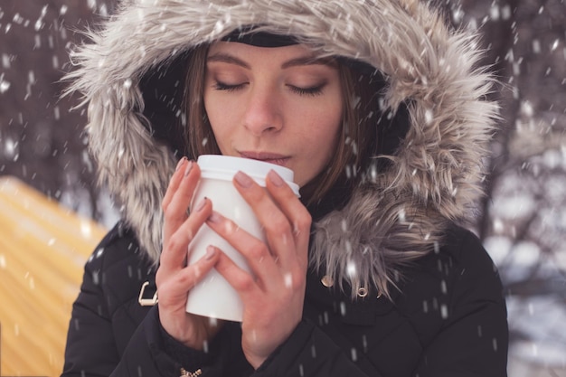 Mulher bebendo café quente ou chá de caneca sob flocos de neve no inverno