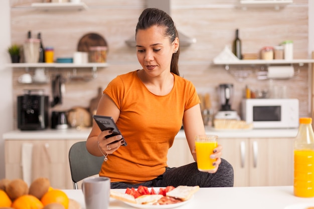 Mulher bebendo suco de laranja fresco pela manhã enquanto navega no smartphone na cozinha