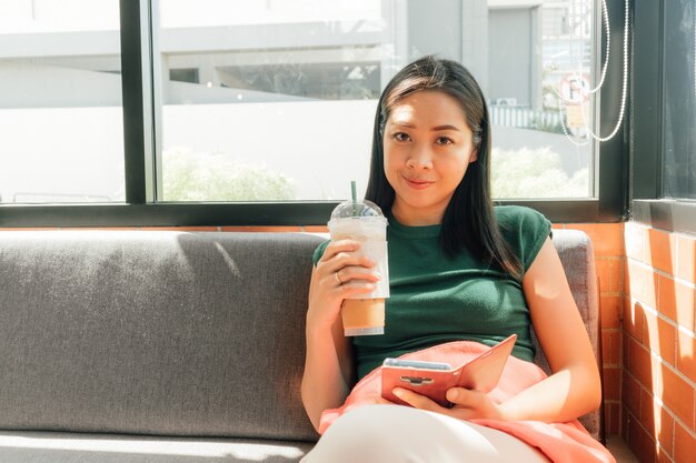 Mulher beber café gelado e usar seu smartphone no sofá no canto do café