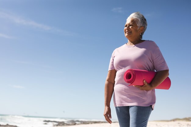 Mulher biracial sênior sorridente segurando um tapete de ioga rosa enquanto olha para a praia contra o céu azul. estilo de vida ativo e fitness.