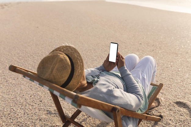 Mulher birracial sênior aposentada usando celular de tela em branco com espaço de cópia na cadeira na praia