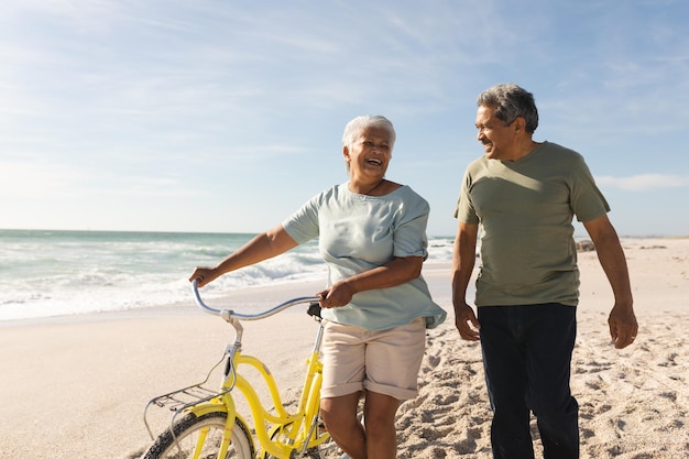 Mulher birracial sênior feliz andando de bicicleta conversando com o homem na praia contra o céu em dia ensolarado