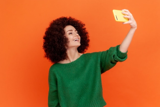 Foto mulher blogueira amigável feliz com penteado afro vestindo suéter verde transmitindo ao vivo falando com seguidores com expressão positiva estúdio interior isolado em fundo laranja